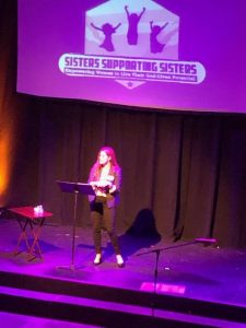 Sisters Supporting Sisters empowerment event on stage with speaker at podium.